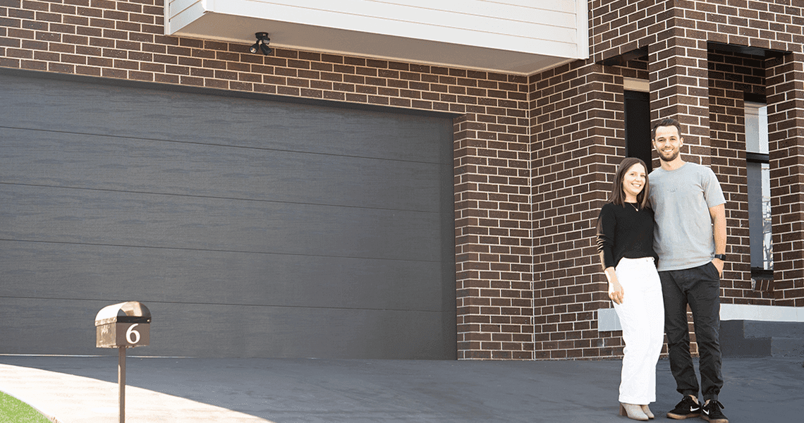 A smiling couple stands arm in arm in front of a modern brick house with a large dark garage door and a mailbox labeled with the number 6.