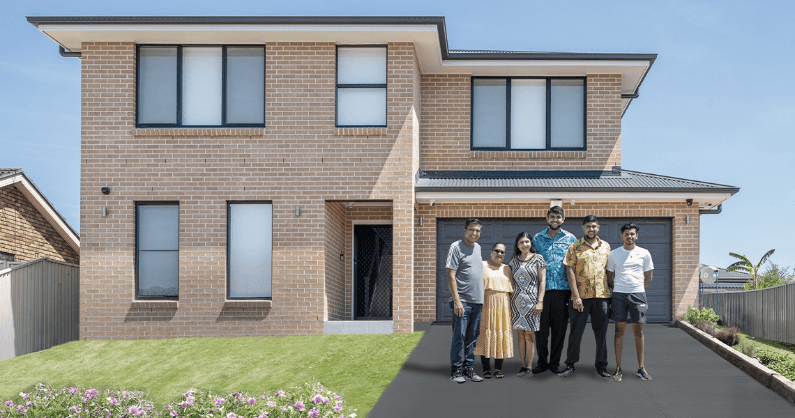 A group of five people stands together in front of a modern two-story brick house with large windows, a double garage, green lawn, and a clear blue sky.