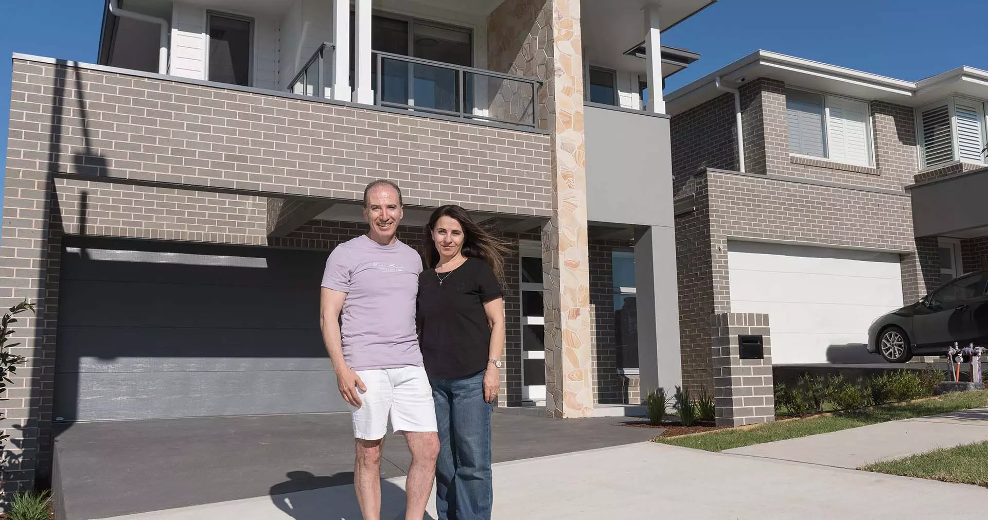 A man and a woman stand smiling together outside a modern, double-story brick house with a large driveway and garage on a sunny day.